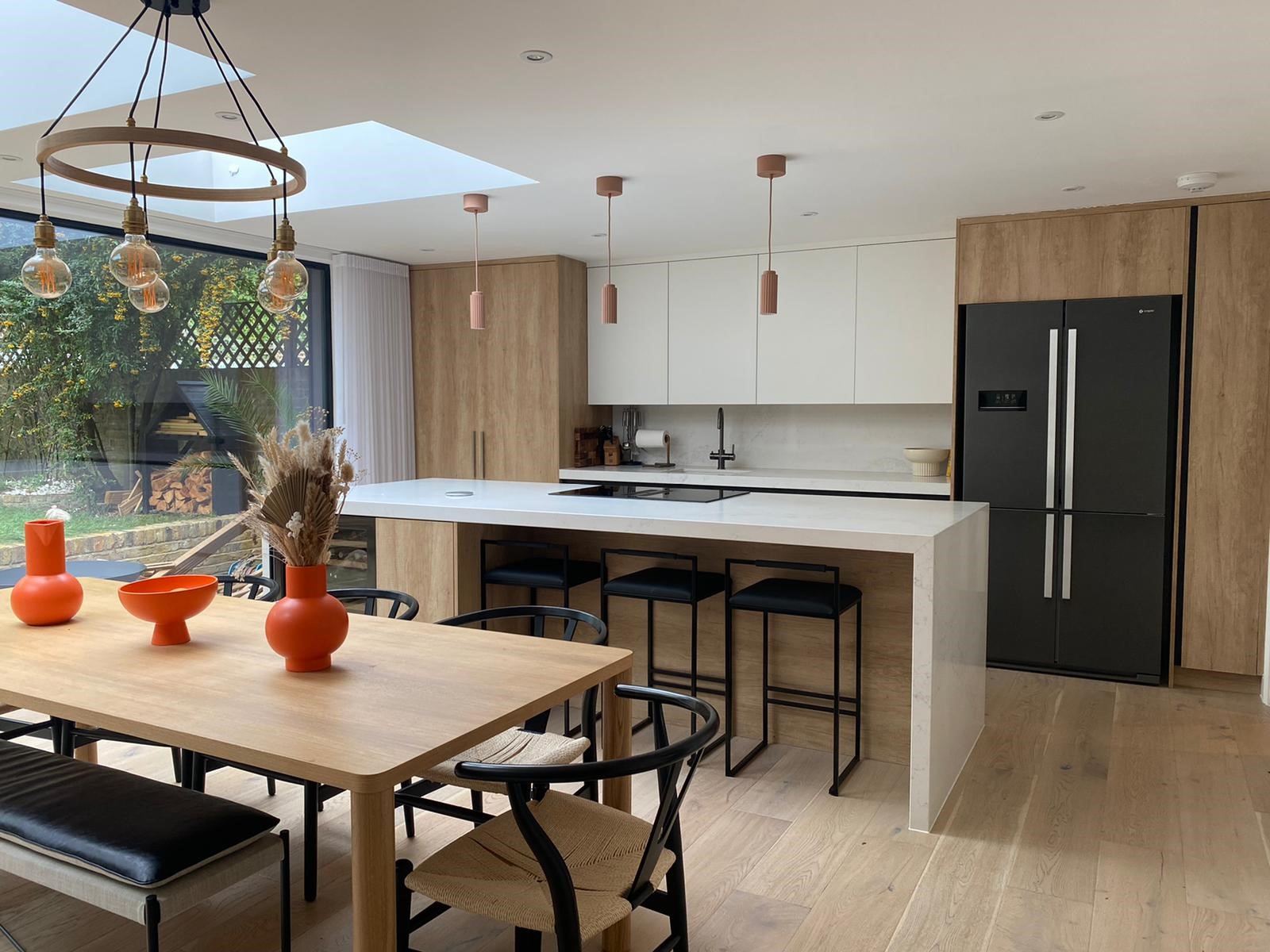 A handleless kitchen mixing warm timber and white units in an open-plan diner, with an island, integrated black fridge and a wall of glazing to the garden.