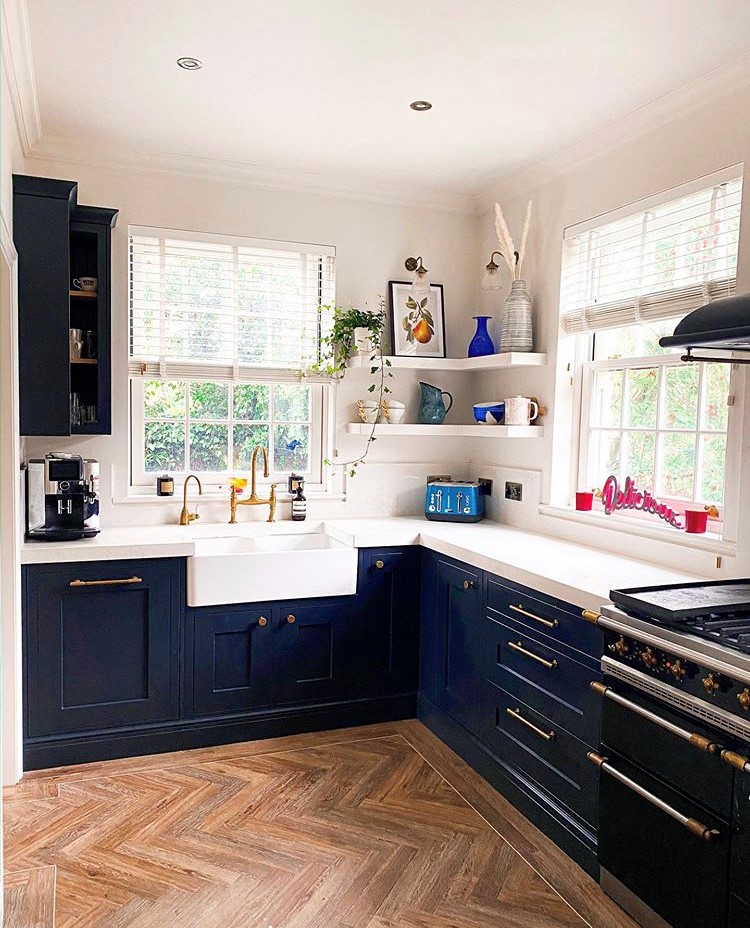 A navy shaker kitchen with a butler sink, brass tapware and floating shelves, wrapped around a bright corner window over herringbone flooring.
